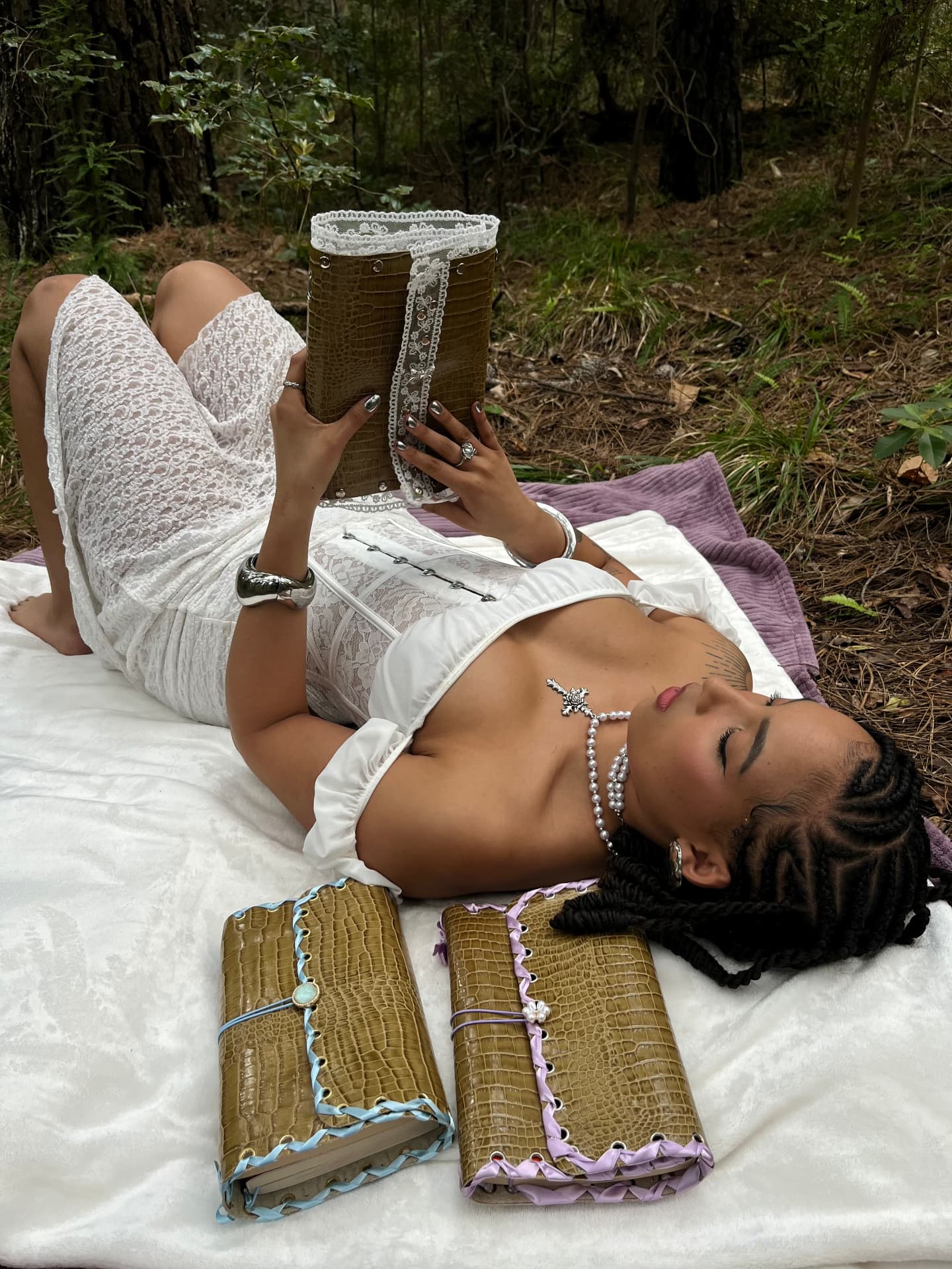 Woman in white lace reading an olive croc leather journal on a forest picnic blanket